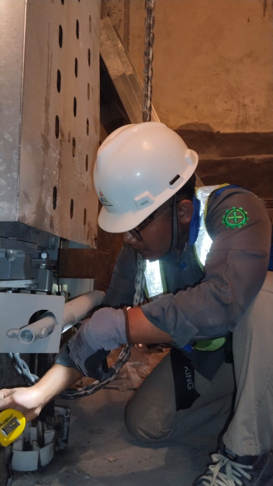 Inspektor ACM berhelm putih berlutut di pit elevator, mengukur jarak runby dengan meteran