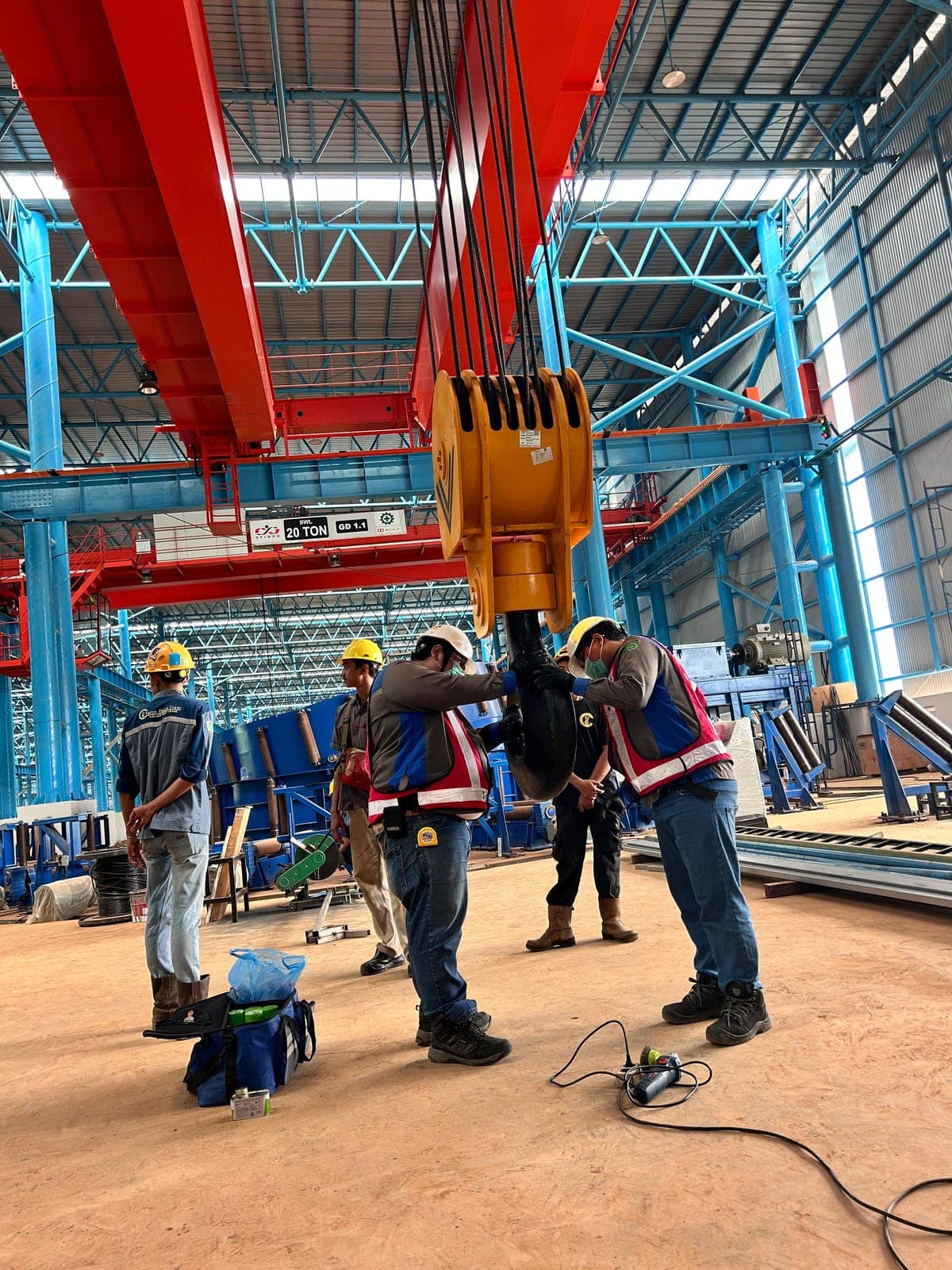 Petugas berhelm dan rompi keselamatan memeriksa hook block besar sebuah overhead crane di dalam pabrik